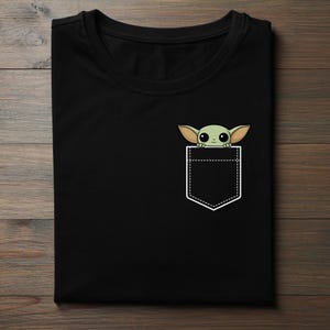 K&ouml;nnte beinhalten: Schwarzes T-Shirt mit einem Taschen-Design, das eine Cartoon-Figur eines gr&uuml;nen Au&szlig;erirdischen zeigt. Der Au&szlig;erirdische hat gro&szlig;e Ohren und Augen, die aus einer wei&szlig;en Taschenkontur hervorschauen. Das T-Shirt ist auf einer Holzoberfl&auml;che gefaltet.