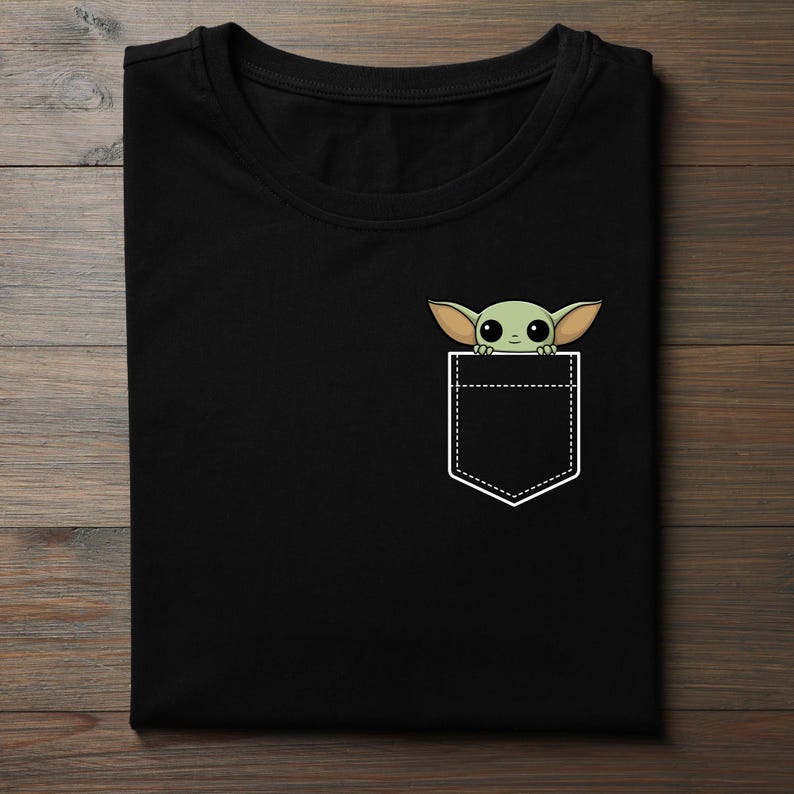 K&ouml;nnte beinhalten: Schwarzes T-Shirt mit einem Taschen-Design, das eine Cartoon-Figur eines gr&uuml;nen Au&szlig;erirdischen zeigt. Der Au&szlig;erirdische hat gro&szlig;e Ohren und Augen, die aus einer wei&szlig;en Taschenkontur hervorschauen. Das T-Shirt ist auf einer Holzoberfl&auml;che gefaltet.