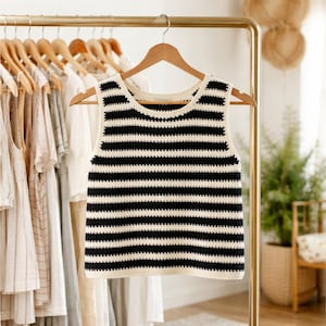 Puede incluir: Un top de crochet a rayas blancas y negras colgado de una percha de madera. El top sin mangas tiene un escote redondo y un patrón de rayas horizontales. Otras prendas de vestir son visibles en una percha en el fondo.