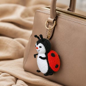 May include: A handmade crochet ladybug keychain with a black head, white body, and red wings with black spots. The ladybug is attached to a gold-colored keyring and is hanging from a beige handbag.