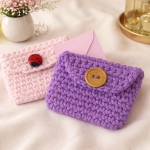 May include: Two handmade crochet envelope-style pouches. One is light pink with a ladybug button, the other is purple with a wooden button. A pink envelope is tucked behind them. The pouches are on a white surface.