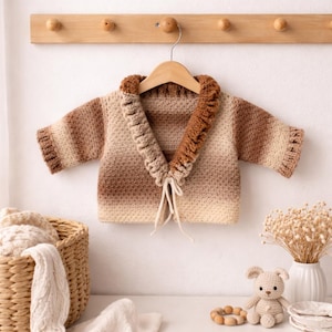 May include: A handmade, crochet baby cardigan in shades of brown and beige hangs on a wooden hanger. The cardigan features a textured stitch and a ruffled collar. A basket of blankets, a crochet bear, and a wooden teether are in the foreground.