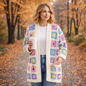May include: A colorful, open-front crochet cardigan with a cream-colored base and square, multi-colored floral patterns. The cardigan has long sleeves and pockets. The person is wearing a white t-shirt and blue jeans.