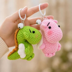 May include: Two crochet keychains: a green dinosaur with an orange spine and a pink hippo. Both have black eyes, white accents, and small bells. The keychains are attached to white rings.