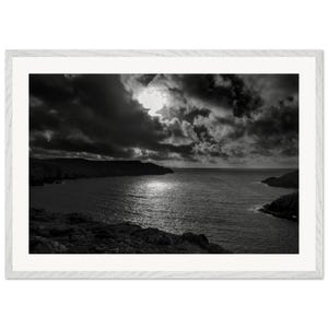 May include: Monochrome framed print depicting a coastal scene. The image shows a dramatic sky with dark clouds above a tranquil sea. The coastline is visible in the foreground, with a bright reflection on the water.