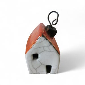 Può includere: Ornamento a forma di casa in ceramica con tetto rosso, pareti bianche e camino nero. L'ornamento ha una piccola finestra e una porta quadrata. Un anello di filo nero è attaccato al camino per appenderlo. La casa ha un aspetto crepato e invecchiato.