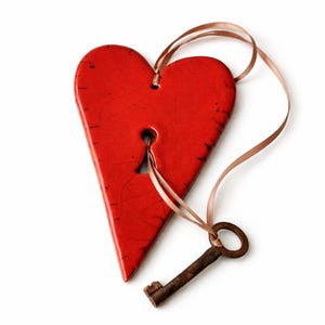 Op de afbeelding: Rood hartvormig ornament met een sleutelgat en een roestige sleutel. Het hart is gemaakt van glanzend rood materiaal en heeft een bruin lint. De sleutel is klein en metaalachtig, met een cirkelvormige handgreep.