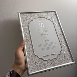 May include: A rectangular silver-framed marriage certificate featuring a white centre panel with the names Bianca and Blake, surrounded by a decorative border with white floral patterns, small pearls, and silver accents on a textured background.