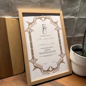 May include: A framed certificate with a light wood frame. The certificate has ornate gold-coloured filigree around the edges and features the names Ethan Alexander Banks and Charlotte Grace Williams. The text reads "This agreement certifies that..."