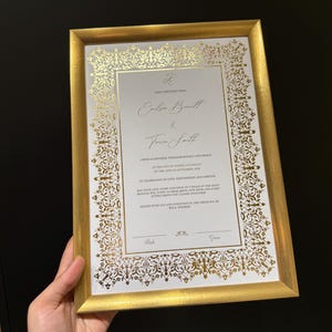 May include: A framed wedding certificate with a gold metallic border and intricate gold filigree patterns. The document features the names Emilsa Bennett and Tricia Smith, with printed text detailing a ceremony in Sydney, Australia, on 19th September 2024.