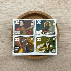 May include: A sheet of four vintage USA postage stamps featuring portraits of Woody Guthrie, Leadbelly, Sonny Terry, and Josh White. Each stamp has a face value of 32 cents. The stamps are on a cork coaster.