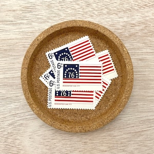 May include: A collection of vintage 6-cent U.S. postage stamps featuring the Bennington Flag of 1777, with red and white stripes and a blue canton with 13 stars. The stamps are arranged in a small, round, cork coaster.