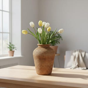 May include: A woven rattan vase filled with white and yellow tulips sits on a light wood table. The vase has a textured, basket-like appearance. The scene is set in a bright room with a window and a plant in the background.