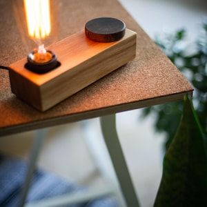 May include: A rectangular light-colored wooden lamp base featuring a single exposed Edison-style light bulb with a warm orange glow and a circular dark wood knob positioned on the top surface of the block for control or adjustment.