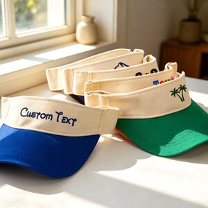 May include: A stack of sun visors with cream-colored headbands and colorful brims in blue, green, and orange. The front headband of the top visor features the embroidered text CUSTOM TEXT in blue, while another visor displays a green palm tree graphic.