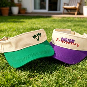 May include: Two sun visors with cream-colored crowns and adjustable straps. One visor features a green brim and an embroidered green palm tree design. The other visor has a purple brim and features the embroidered text CUSTOM Embroidery.