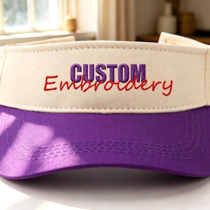 Könnte beinhalten: Ein zweifarbiger Sonnenschutz mit cremefarbenem Vorderteil und violettem Schirm. Auf dem Vorderteil befindet sich der Text "CUSTOM" in violetten Blockbuchstaben über dem Wort "Embroidery" in roter Schreibschrift, beides gestickt.