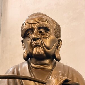 Peut inclure: Statue en bronze d'un homme âgé au regard serein. La statue présente des traits de visage détaillés, notamment des rides et un léger sourire. L'homme tient un long objet plat, peut-être un bâton ou un instrument d'écriture. La statue est placée sur un fond neutre.