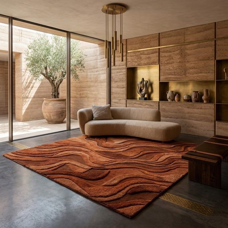 May include: A contemporary living room with a rust-coloured rug featuring a wave design. A curved beige sofa and a wooden bench are also present. The room has a large window with an olive tree in a terracotta pot, and a gold light fixture.
