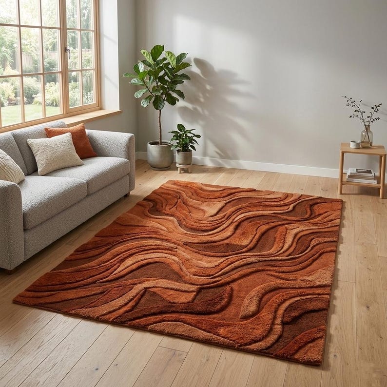 May include: A rectangular rug with a textured, layered design in shades of orange and brown. The rug's wavy pattern creates a 3D effect. It's placed on a wooden floor in a living room, adding warmth and texture to the space.