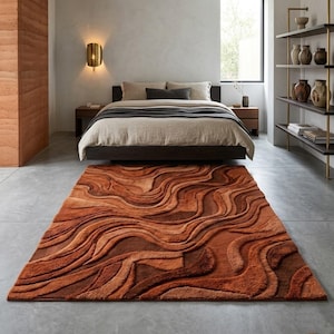 May include: A large, rectangular rug with a textured, wave-like pattern in shades of orange and brown. The rug is placed in a bedroom setting, in front of a dark wooden bed frame. The room has a neutral colour palette.