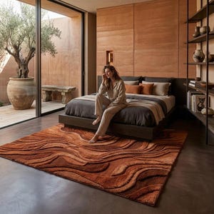May include: A bedroom with a rust-coloured rug with a wave design. The rug is on a concrete floor in front of a bed with dark bedding and pillows. A person sits on the bed's edge. The room has a large window and a neutral colour scheme.