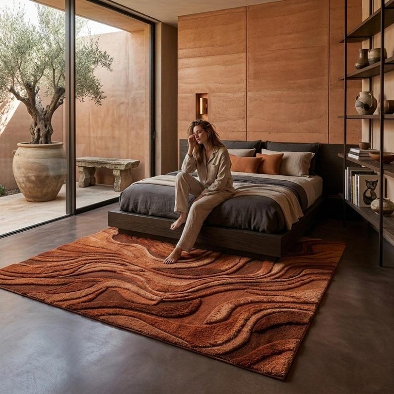 May include: A bedroom with a rust-coloured rug with a wave design. The rug is on a concrete floor in front of a bed with dark bedding and pillows. A person sits on the bed's edge. The room has a large window and a neutral colour scheme.