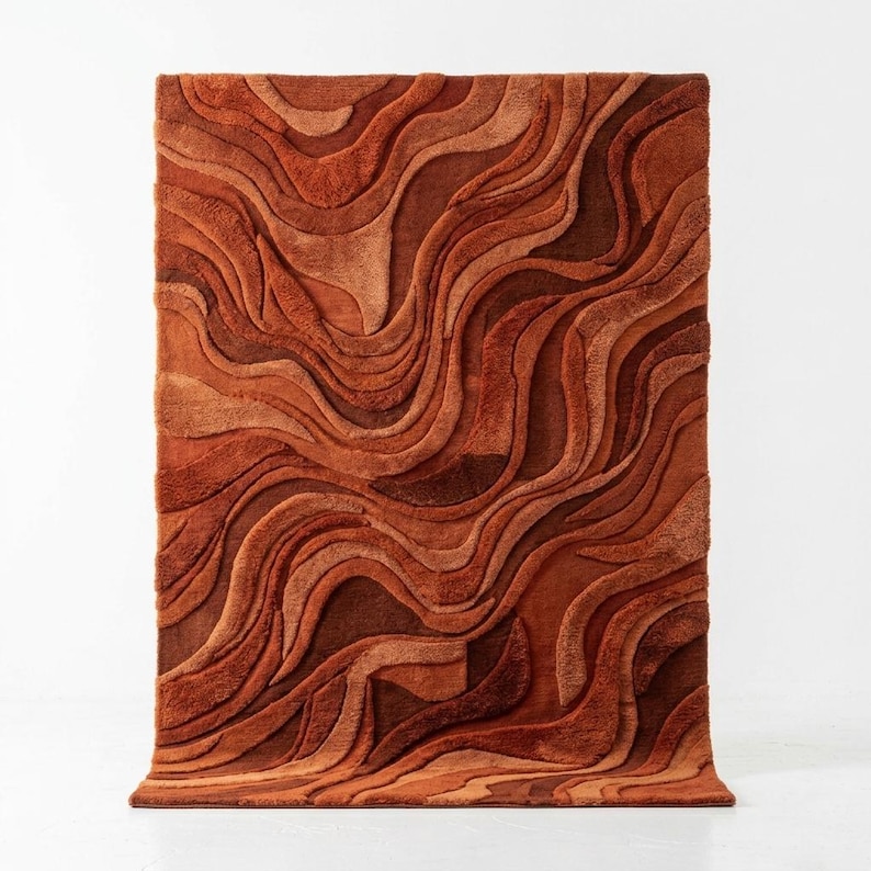 May include: A rectangular rug with a textured surface, in shades of orange and brown. The rug's design features a wavy, layered pattern, creating a three-dimensional effect. The colour palette ranges from light orange to deep brown, adding depth to the floor covering.