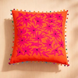 Puede incluir: Cojín cuadrado naranja con diseño floral rosa bordado. El cojín está adornado con un borde de pompones de colores. Un elemento decorativo brillante y alegre para cualquier habitación.