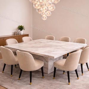 Puede incluir: Un comedor moderno con una gran mesa cuadrada de mármol y ocho sillas beige. Una lámpara de araña redonda con múltiples globos de cristal cuelga sobre la mesa. La habitación está decorada con tonos neutros y una alfombra gris claro. La mesa mide unos 200 cm x 200 cm.