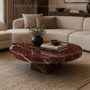 May include: A circular, red marble coffee table with white veining, set on a square base. The table is in a living room with a beige sofa, a rug, and decorative items. The table has books and a bowl on top.