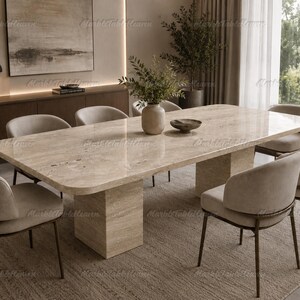 Pode incluir: Uma mesa de jantar retangular com um tampo de pedra travertino bege polido e duas bases de pedestal correspondentes em forma de bloco. A mesa é cercada por seis cadeiras estofadas com encostos curvos e pernas finas de metal escuro.