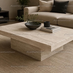 Pode incluir: Uma mesa de centro quadrada feita de pedra travertino bege com uma textura porosa natural. A mesa apresenta um tampo espesso e plano apoiado por uma base retangular larga e sólida, criando uma estrutura baixa e compacta.