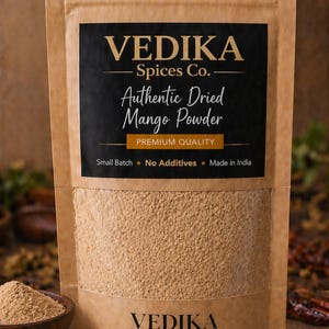 May include: A brown paper bag of Vedika brand Authentic Dried Mango Powder. The bag has a black label with gold text. The text on the label reads "Authentic Dried Mango Powder" and "Premium Quality". A small wooden bowl of mango powder is in the foreground.