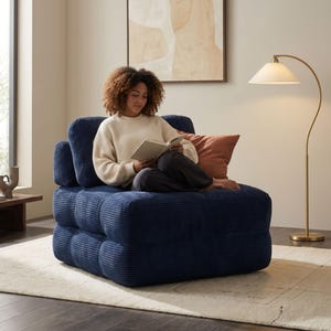 May include: A navy blue corduroy armchair with a square, modular design. The chair has a low profile and a textured surface. A person is sitting on the chair, reading a book. A peach-colored pillow rests on the chair.