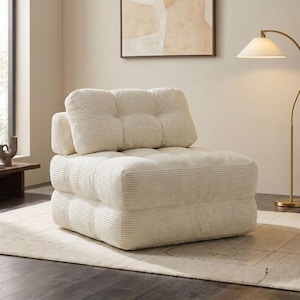 May include: Cream-colored corduroy armchair with a square, modular design. The chair features a tufted back and seat, and is placed on a neutral-toned rug. A floor lamp and artwork are in the background.