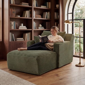 May include: A green corduroy chaise lounge with a matching armchair. A person is reading a book titled "The Silent Hours" while sitting on the chaise lounge. The background features a wooden bookshelf and a brass floor lamp.