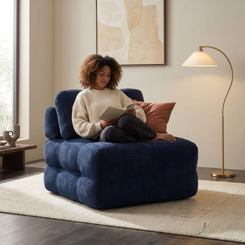 May include: A navy blue corduroy armchair with a square, modular design. The chair has a low profile and a textured surface. A person is sitting on the chair, reading a book. A peach-colored pillow rests on the chair.