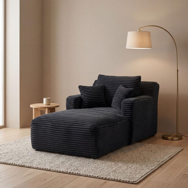 May include: A dark gray corduroy chaise lounge with two small square pillows and a large back cushion. A wooden side table with a white mug sits to the left of the lounge. A gold floor lamp with a beige lampshade is to the right.