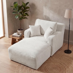 May include: A white corduroy chaise lounge with matching pillows. The lounge has a wide seat and a backrest with two additional pillows. The room also includes a small wooden side table, a floor lamp, and a potted plant.