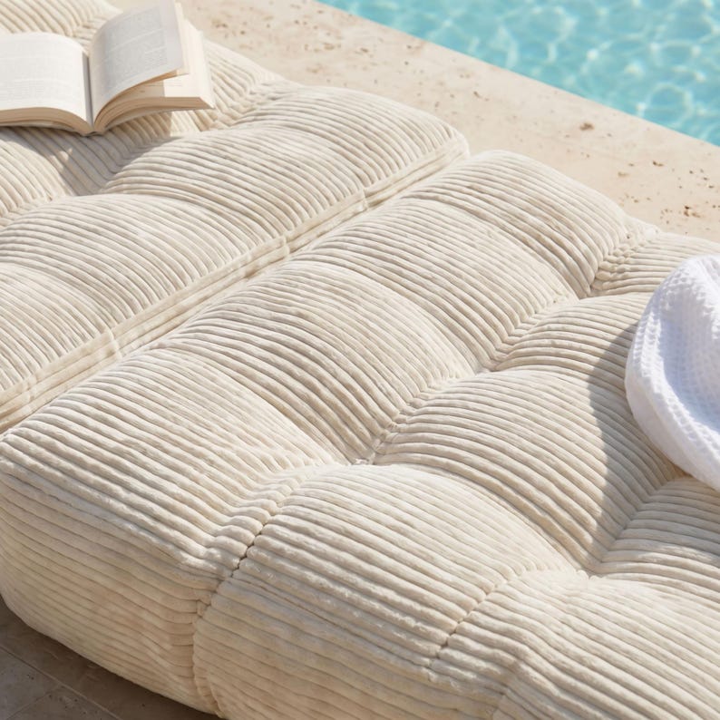 May include: Cream-colored corduroy outdoor cushions with a tufted design. An open book rests on one cushion, and a white sun hat is placed nearby. The cushions are positioned near a pool.
