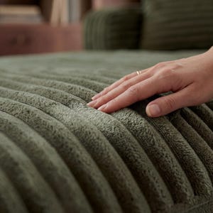 May include: Close-up of a sage green, corduroy-textured fabric. The fabric has a ribbed pattern and a soft, plush appearance. A hand with a gold ring gently touches the fabric, highlighting its texture and color.