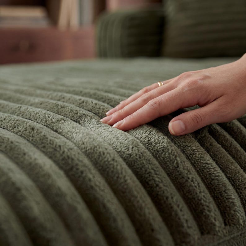 May include: Close-up of a sage green, corduroy-textured fabric. The fabric has a ribbed pattern and a soft, plush appearance. A hand with a gold ring gently touches the fabric, highlighting its texture and color.