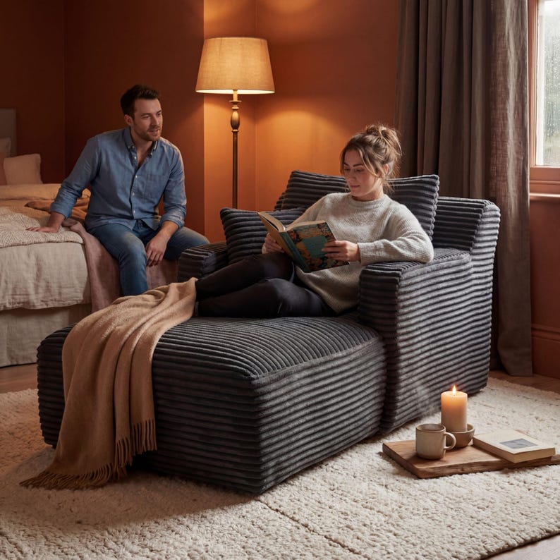 May include: A gray corduroy chaise lounge with a matching pillow and armrests. A person is reading a book while sitting on the chaise lounge. A tan blanket is draped over the footrest. A wooden tray holds a candle, mug, and book.