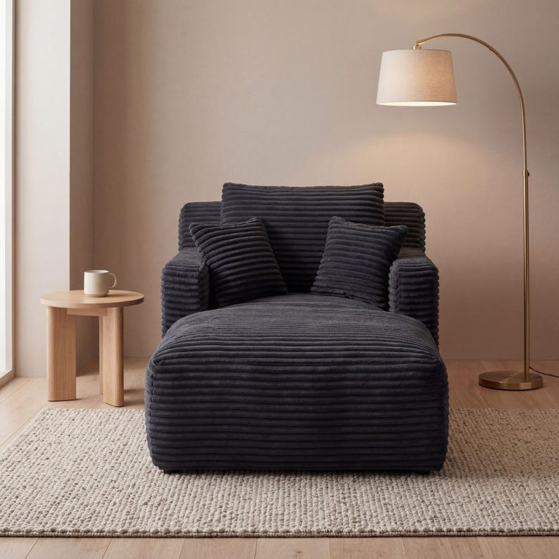 May include: A dark gray corduroy lounge chair with matching pillows and a footrest. The chair is next to a small wooden side table with a white mug and a gold floor lamp with a beige lampshade.