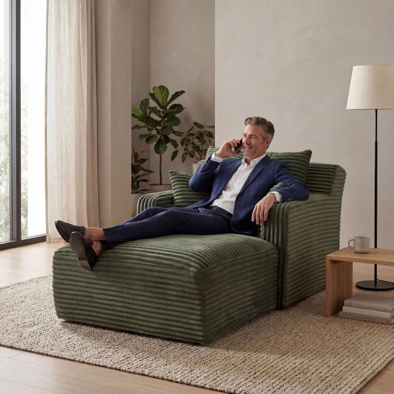 May include: A green corduroy armchair with an attached ottoman. The chair has a boxy shape and is in a living room setting. The person is wearing a blue suit and is on the phone. The room has neutral tones and a natural fiber rug.