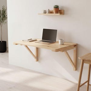 Könnte beinhalten: Ein helles, an der Wand montiertes Holzschreibtisch mit Laptop, Notizbuch und Becher. Ein kleines Holzregal darüber enthält eine Topfpflanze und dekorative Gegenstände. Der Schreibtisch wird von abgewinkelten Holzhalterungen getragen.