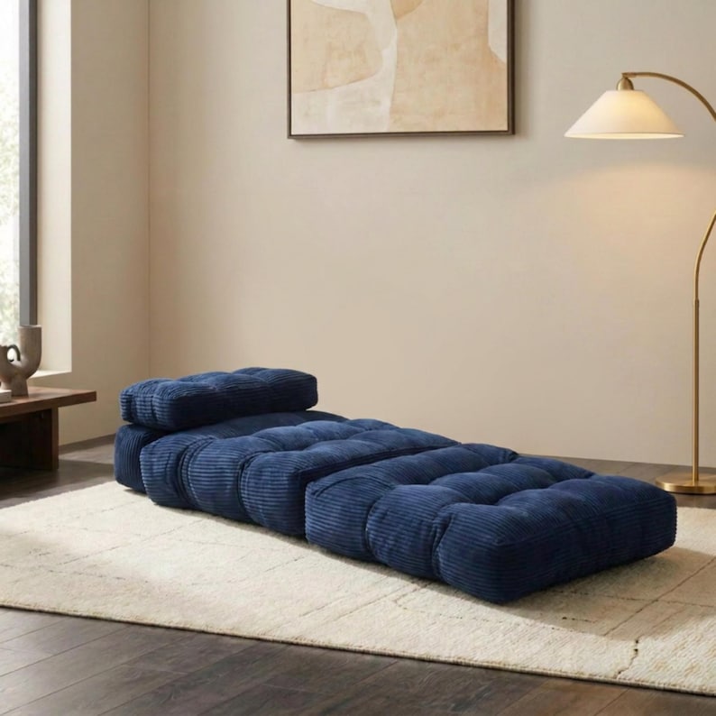 May include: A navy blue corduroy chaise lounge with a matching headrest. The lounge is composed of several square, cushioned sections. It sits on a cream-colored rug in a room with a neutral color palette.