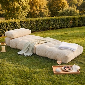 May include: A beige corduroy outdoor daybed with a matching pillow, a light green blanket, and a white towel. A wooden tray holds a glass of wine, figs, and a napkin, set on a grassy lawn.
