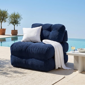May include: A navy blue corduroy outdoor lounge chair with a white pillow and blanket. The chair is near a pool with olive trees in the background. A small white side table holds a glass of water and a bowl.